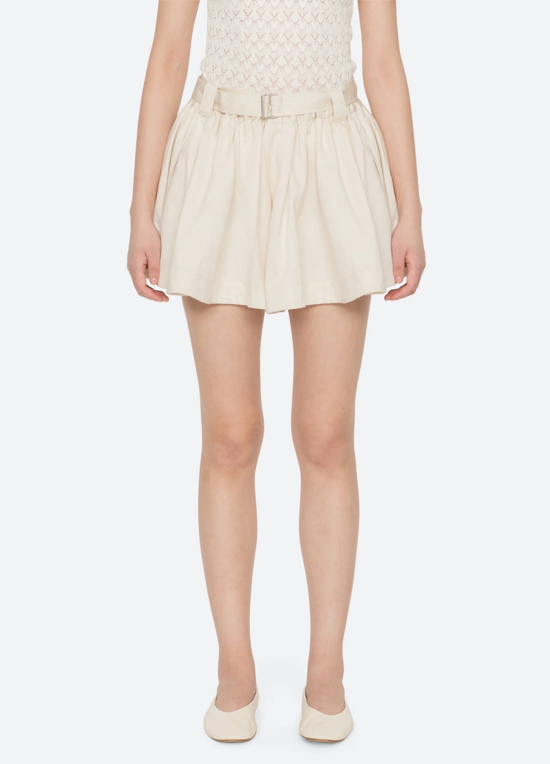 A person wears the Sea New York Angelina Suiting Flare Short—high-waisted, pleated, with an elastic waistband and belt—paired with a cream textured sleeveless top and matching cream flats, standing against a plain light background.