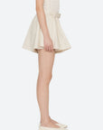A person stands sideways against a plain light background, wearing Sea New York’s Angelina Suiting Flare Short in beige with an elasticized waistband and a sleeveless top, paired with cream-colored flats.