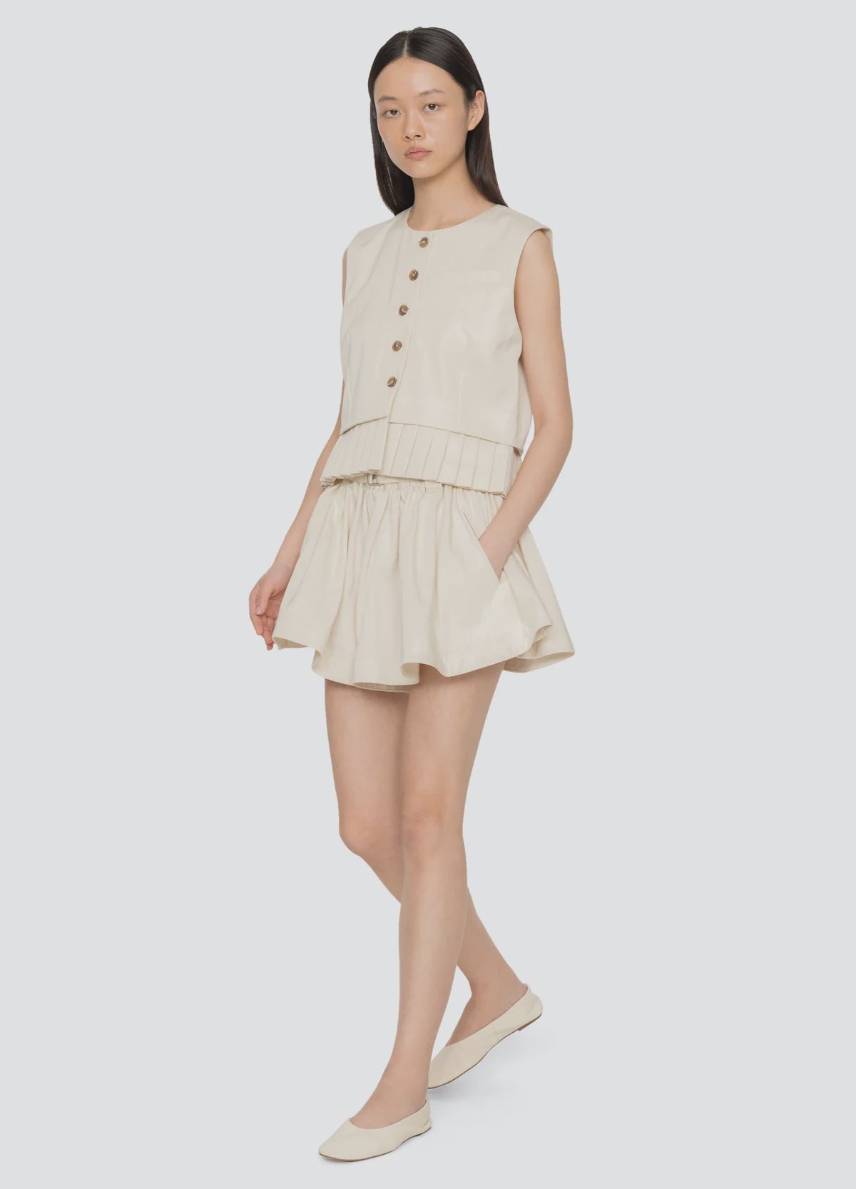 A woman with straight long hair stands against a plain background wearing the Sea New York Angelina Suiting Flare Short, a sleeveless beige button-up top, and matching beige flats. The shorts feature pleats and an elasticized waistband.