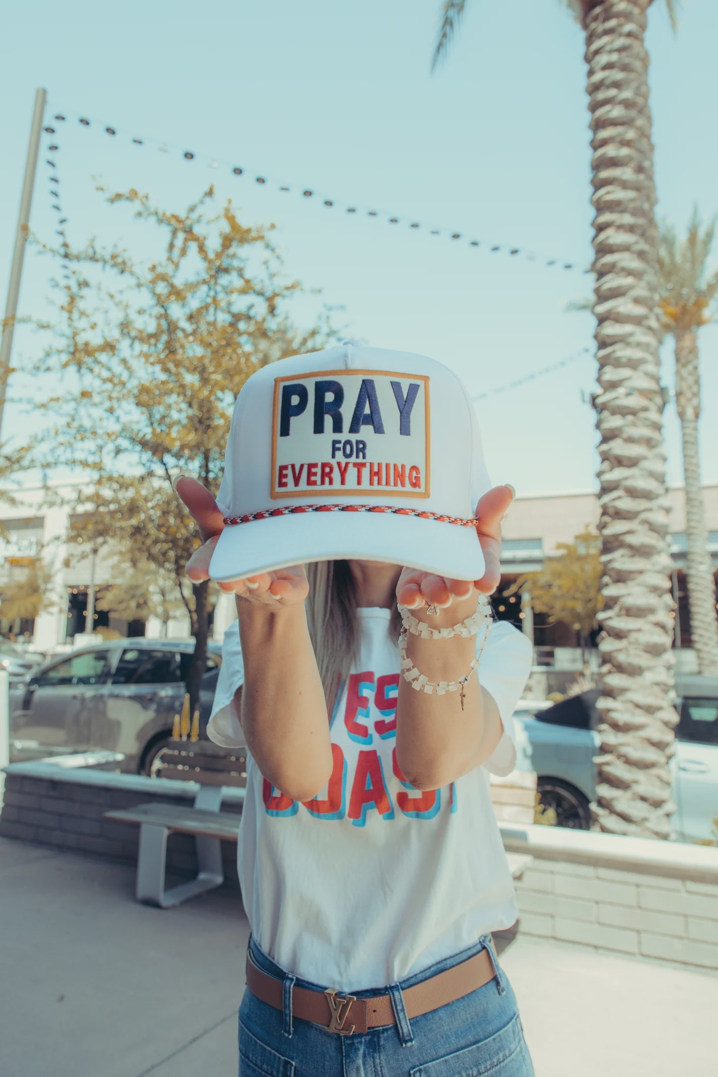 A person with their face partially hidden by a **PRAY FOR EVERYTHING hat** from **Pray for Everything** stands outside. Wearing a white T-shirt and jeans, they are surrounded by palm trees and cars in the background, creating a relaxed atmosphere.