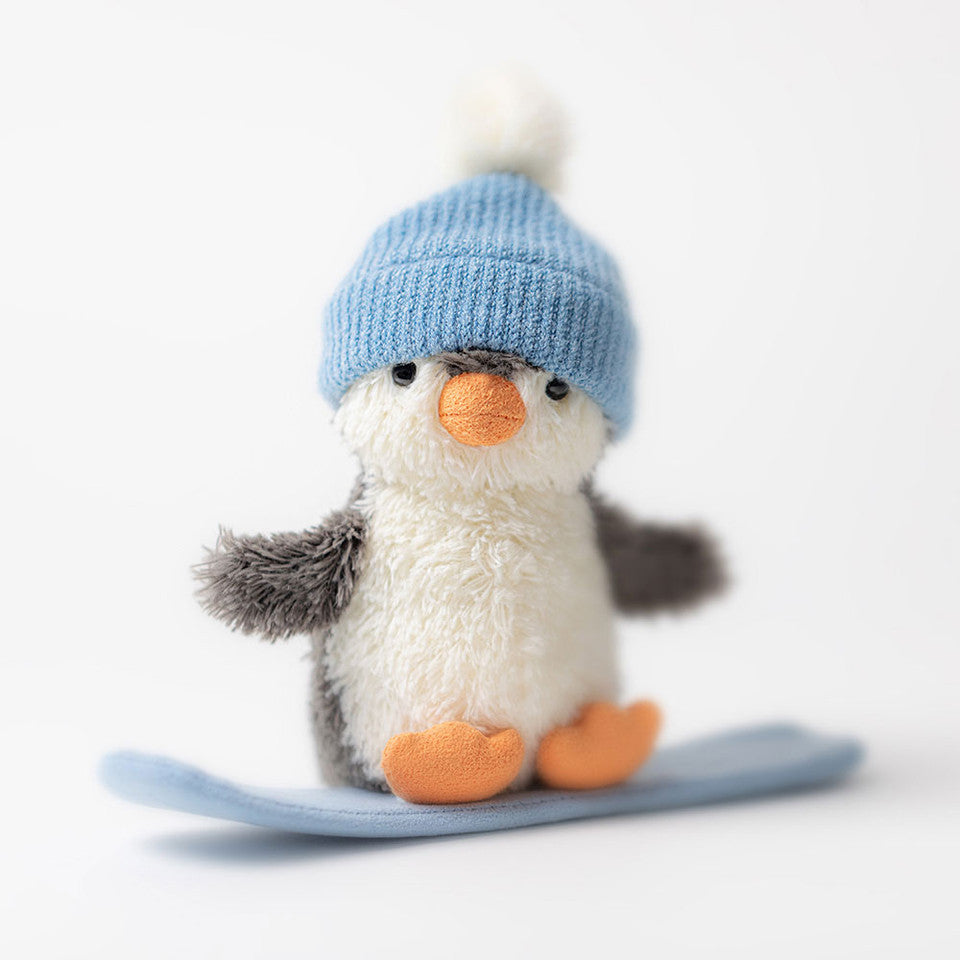 The Jelly Cat Inc. Peanut Penguin Snowboarding plush toy wears a light blue knit hat and stands on a small blue snowboard against a plain white background.