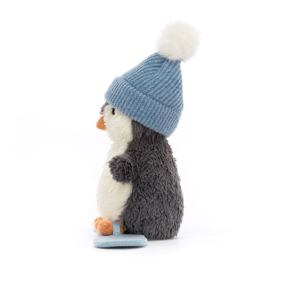 The Jelly Cat Inc. Peanut Penguin Snowboarding plush sits upright in a blue knit hat with white pom-pom, featuring a white belly, gray back, and orange beak and feet, shown in profile on a white background.