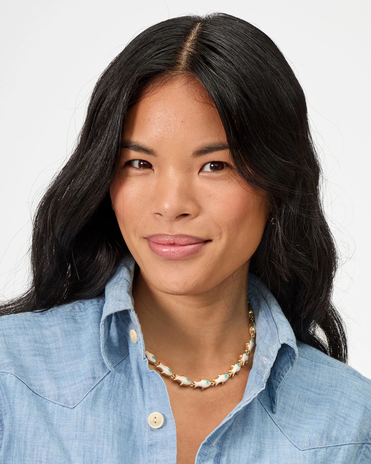 A woman with long black hair and medium skin tone smiles gently, wearing a light blue button-up shirt and the Clare Vivier Pearl Sardine Necklace in gold plated brass, set against a plain white background.