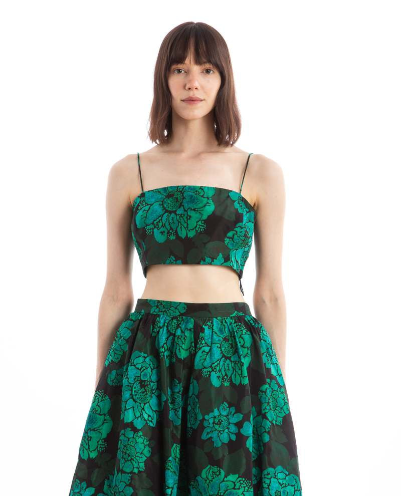 A woman with shoulder-length hair showcases a bold floral outfit by Rhode. The Penny Top and matching skirt, both in black, feature eye-catching large green flowers. She exudes confidence against a plain white background.