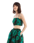 Dressed in a chic Rhode Penny Top paired with a matching high-waisted skirt, the ensemble features an elegant green and black floral pattern. The sleeveless crop top enhances the look as they confidently pose against a plain white background.