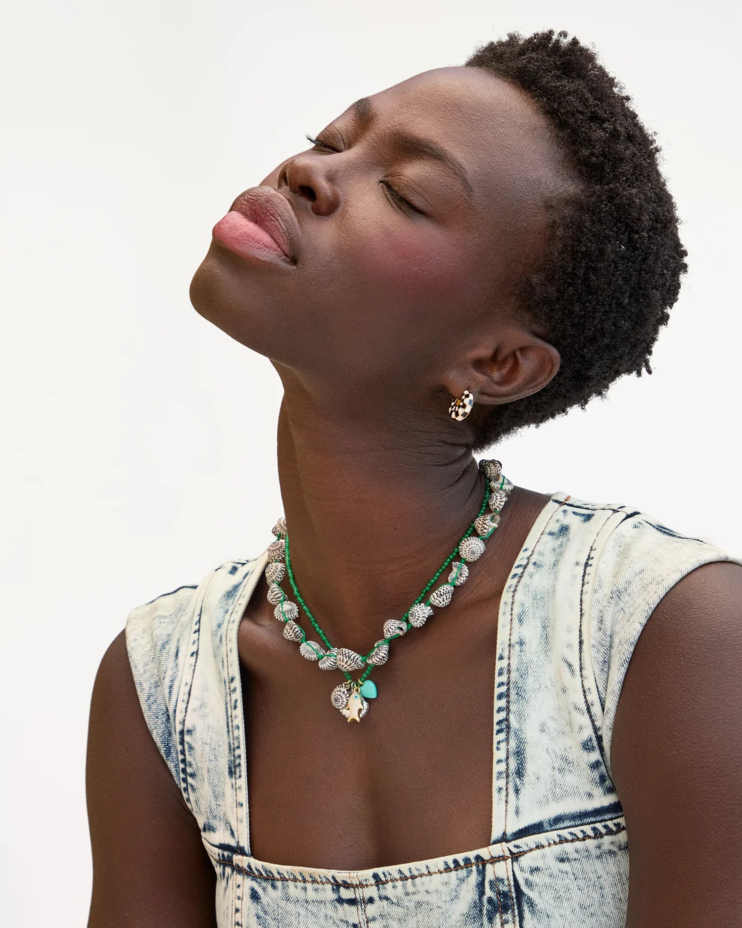 A woman with short curly hair tilts her head back, eyes closed, wearing a sleeveless acid-wash denim top and the Clare Vivier Checker Shell Necklace, along with layered green and silver necklaces, gold stud earrings, against a white background.