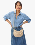 A person in a blue denim shirt and skirt stands with hands on hips, showcasing the Clare Vivier Petit Moyen Messenger. The beige crossbody bag features a perforated design and an adjustable strap against a plain white background.