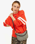 A person with short hair wearing an oversized orange sweater with white stripes on the sleeves and a leopard print skirt poses with Clare Vivier's Petit Moyen Woven Checker bag, featuring handwoven leather crossbody straps, slung over their shoulder. They are gazing to the side with a neutral expression.