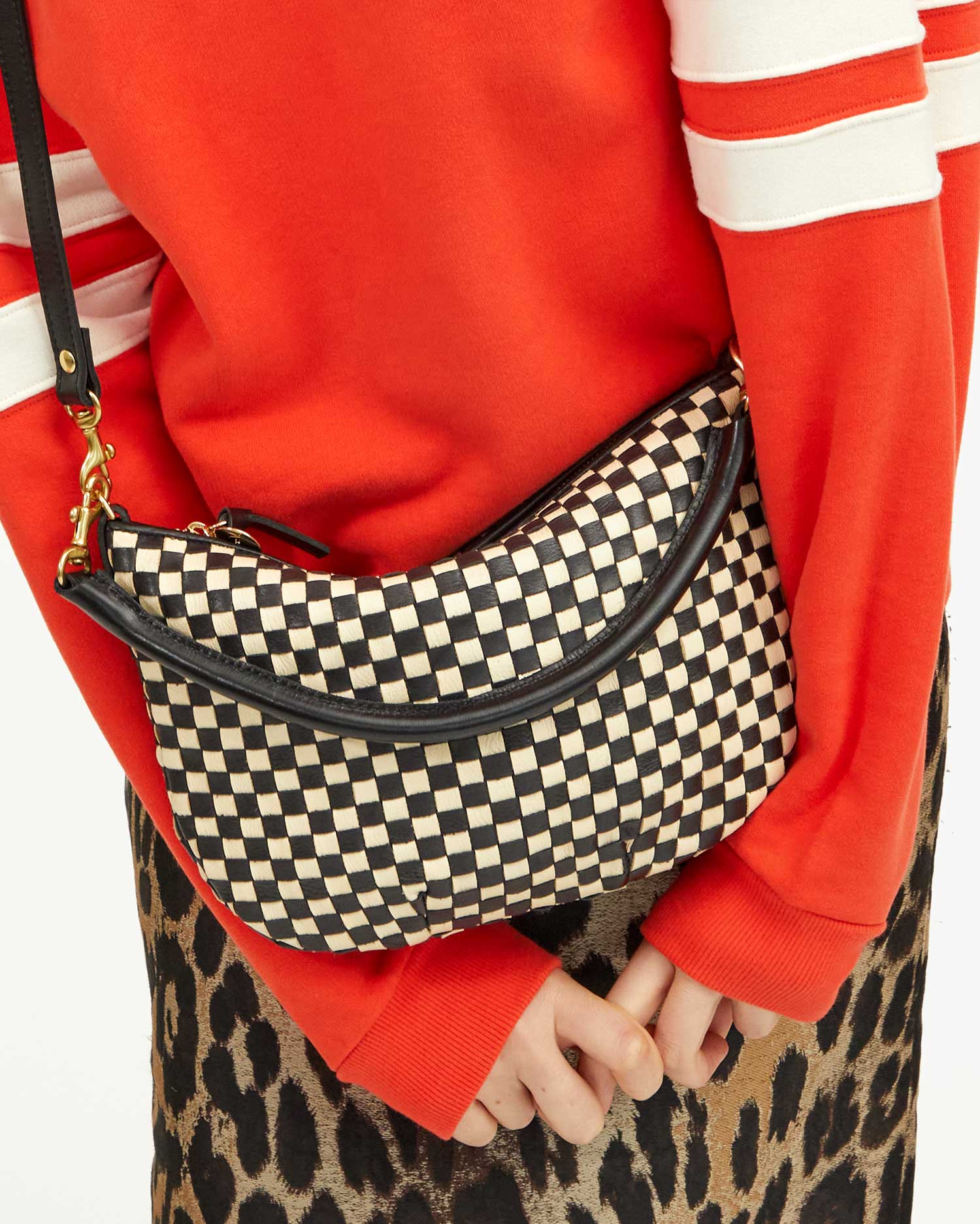 A person dressed in a red sweater with white stripes on the sleeves is holding a Clare Vivier Petit Moyen Woven Checker mini messenger behind their back. The individual is also wearing a leopard print skirt.