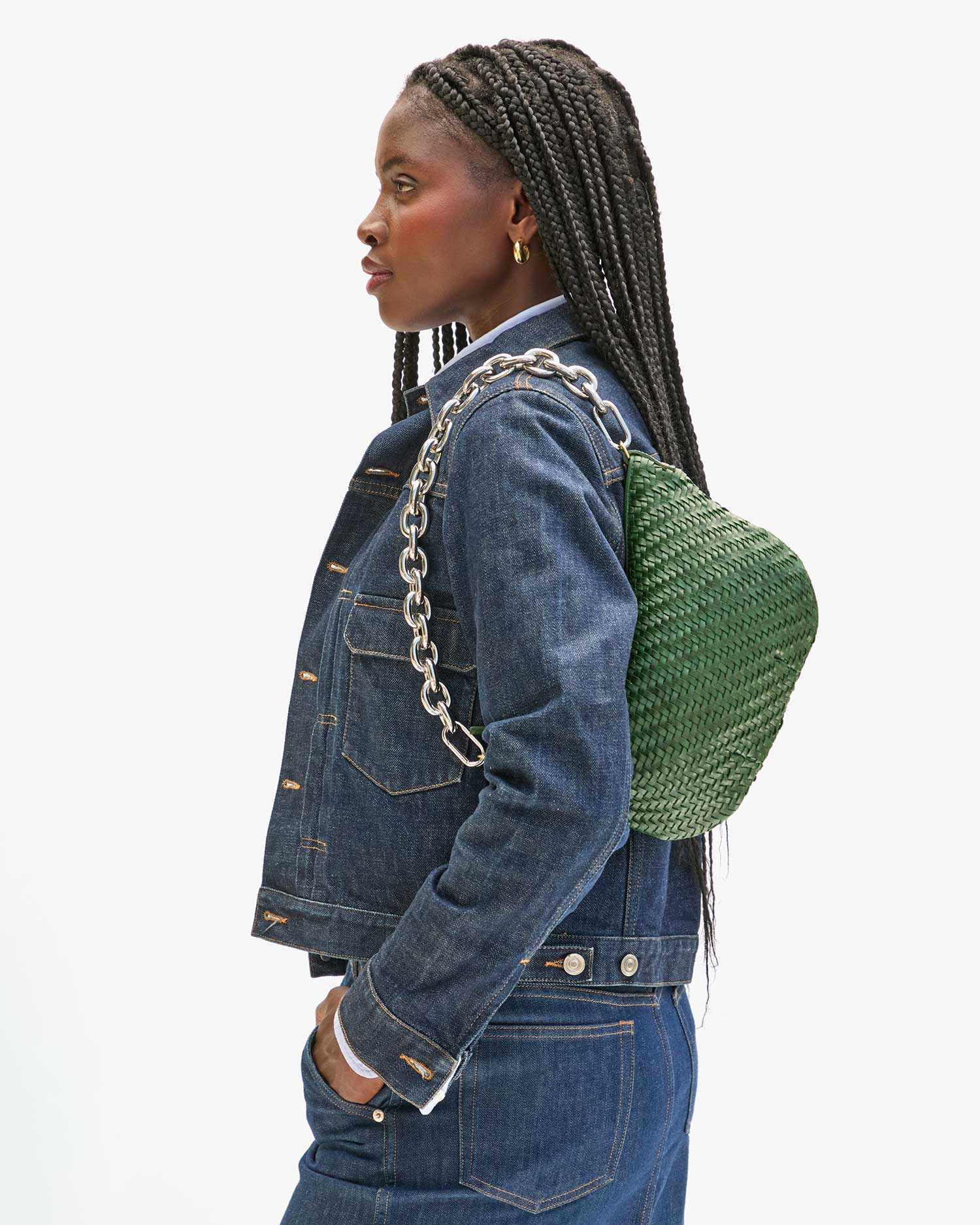 A person in a denim jacket and jeans with braided hair looks to the side, carrying a green woven bag featuring Clare Vivier's eye-catching 18-inch gold & silver mix Shortie Strap over one shoulder. The background is plain white.