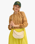 A person wearing a brown blouse, yellow skirt, and green cap with "Déjà Vu" written on it holds the Clare Vivier Petite Moyen S24, a handwoven leather handbag featuring crossbody straps. They stand against a plain background with a relaxed expression.