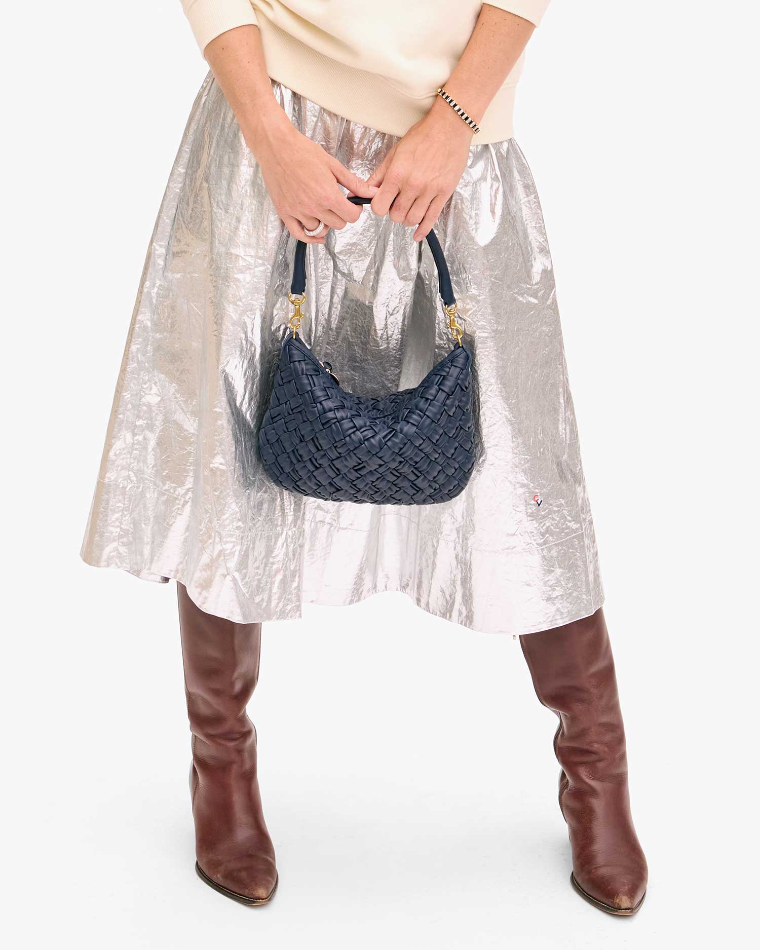 A person holding the Petite Moyen handbag in dark blue with chrome studs from Clare Vivier stands against a white background. They are wearing a metallic silver midi skirt, a cream sweater, and brown knee-high boots featuring zipper closures. The image highlights the outfit without revealing the person&#39;s face.