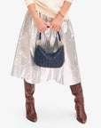 A person holding the Petite Moyen handbag in dark blue with chrome studs from Clare Vivier stands against a white background. They are wearing a metallic silver midi skirt, a cream sweater, and brown knee-high boots featuring zipper closures. The image highlights the outfit without revealing the person's face.