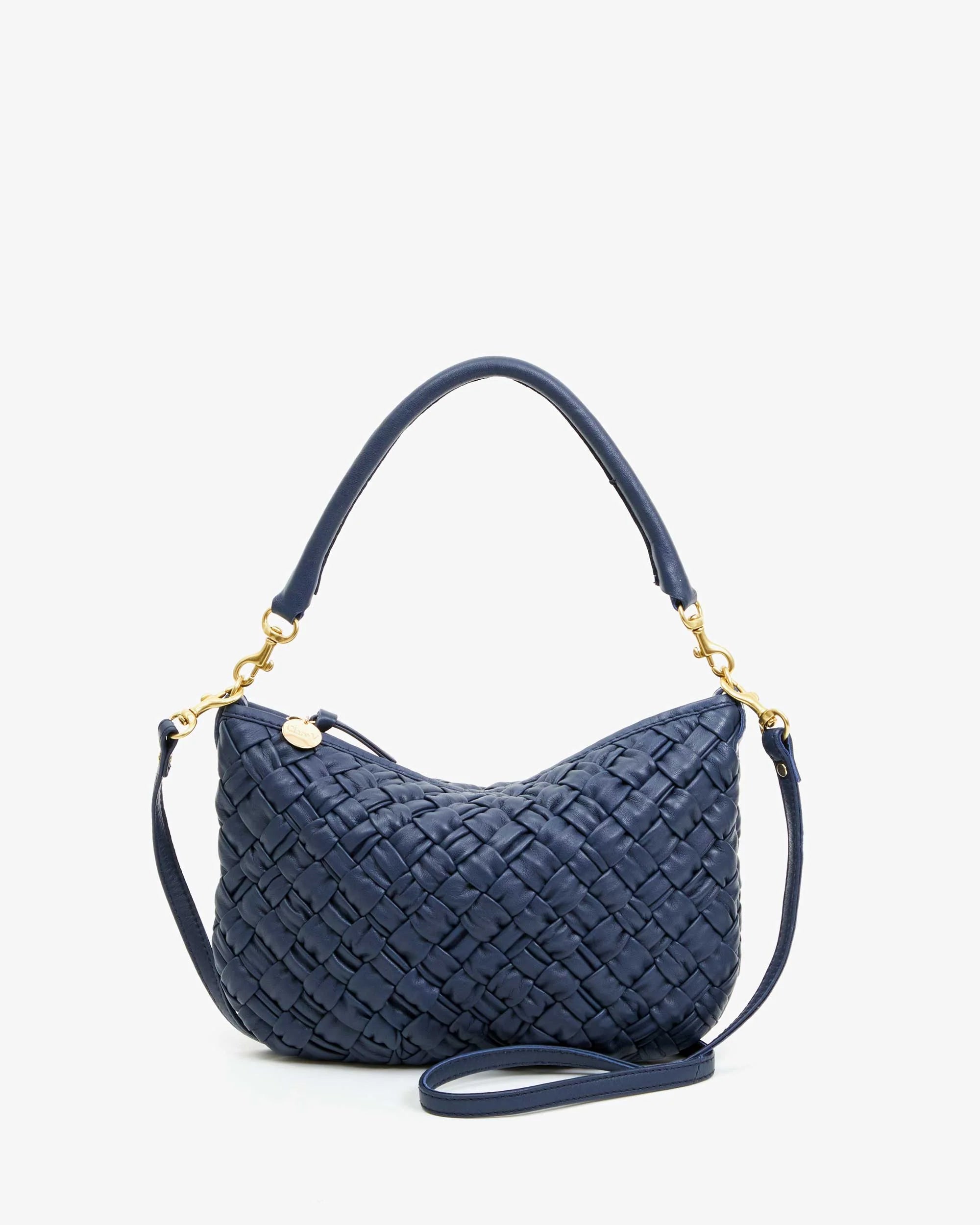 Featuring a suede finish, gold hardware, and chrome studs, the Petite Moyen handbag from Clare Vivier is a stylish navy blue woven accessory with a single top handle and a detachable crossbody strap. It boasts a secure zipper closure, all beautifully displayed against a plain white background.