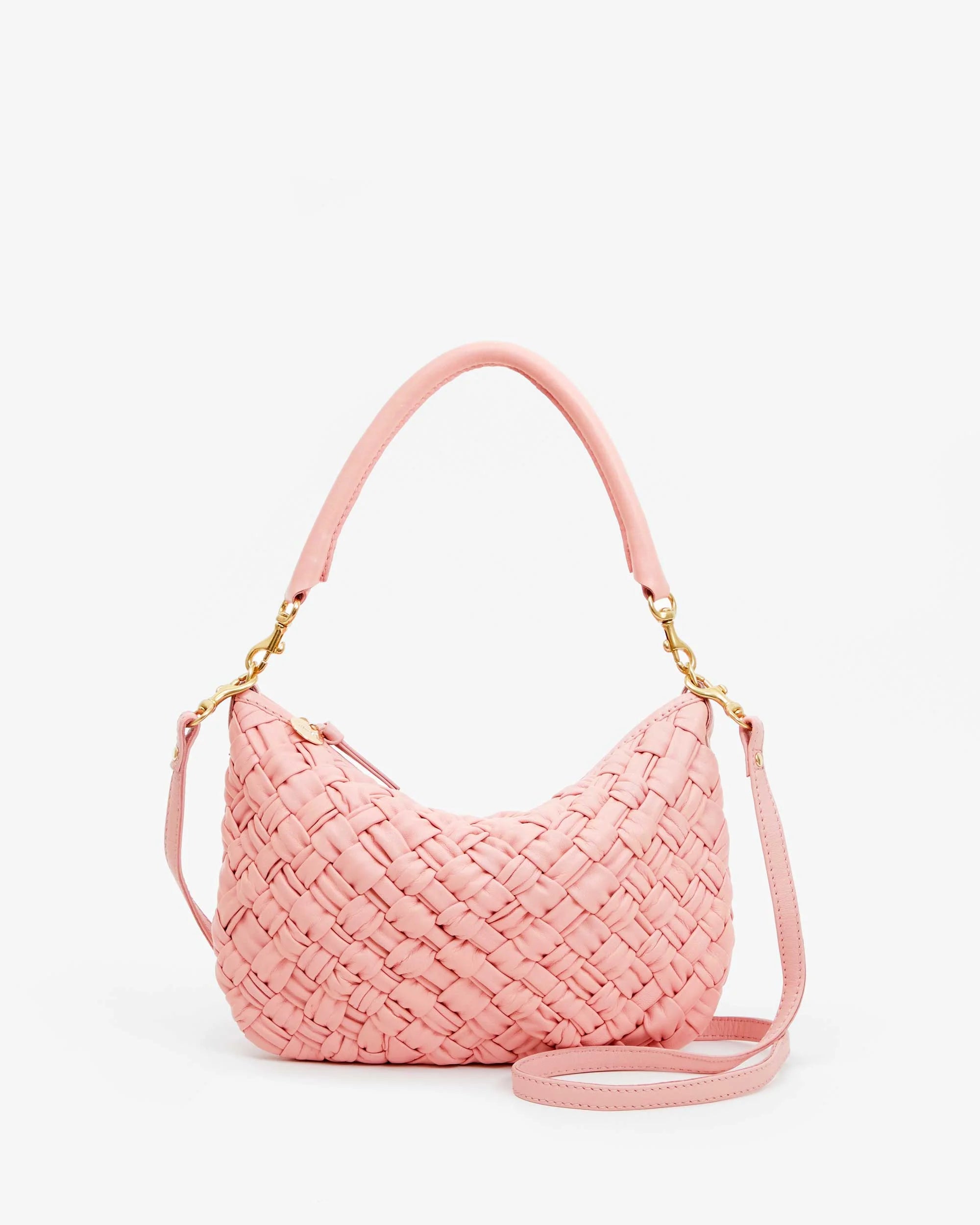 The Petite Moyen by Clare Vivier is a pink, handwoven leather handbag with a short handle and detachable crossbody strap. Featuring gold-tone hardware and a crescent shape, its woven texture adds style and trendiness against a plain white background.