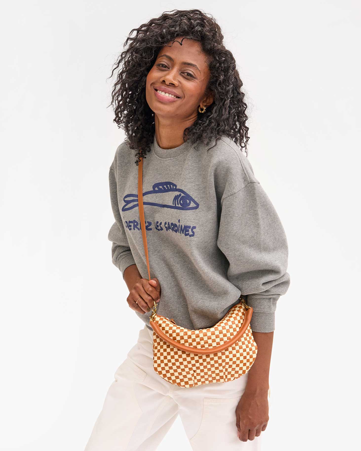 A person with curly hair smiles, wearing a gray sweatshirt with a fish graphic and text, paired with white pants. They hold the Petit Moyen Woven Checker mini messenger bag by Clare Vivier, which features a brown and white checker pattern and comes with a crossbody strap.