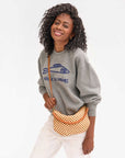 A person with curly hair smiles, wearing a gray sweatshirt with a fish graphic and text, paired with white pants. They hold the Petit Moyen Woven Checker mini messenger bag by Clare Vivier, which features a brown and white checker pattern and comes with a crossbody strap.