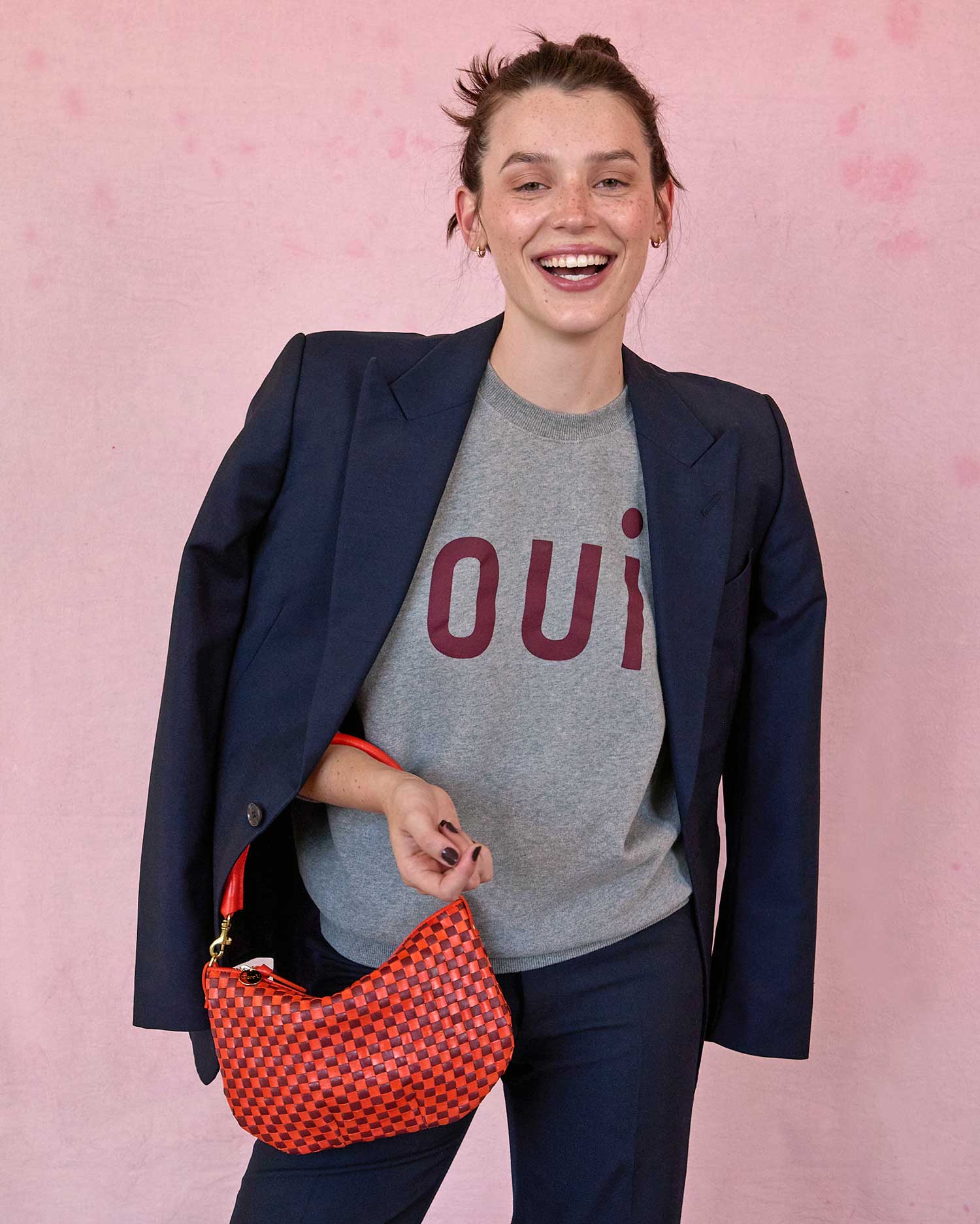 A smiling person stands against a pink background, wearing a gray shirt with "Oui" in burgundy letters, a black blazer, and holds the Clare Vivier Petit Moyen Woven Checker handbag featuring a handwoven leather design and stylish crossbody strap.