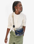 A woman with braided hair wears a cream blouse and olive green pants. She carries the Clare Vivier Petite Moyen S24, a blue mini messenger bag featuring handwoven leather and a wide crossbody strap over her shoulder. She looks to the side against a plain background.