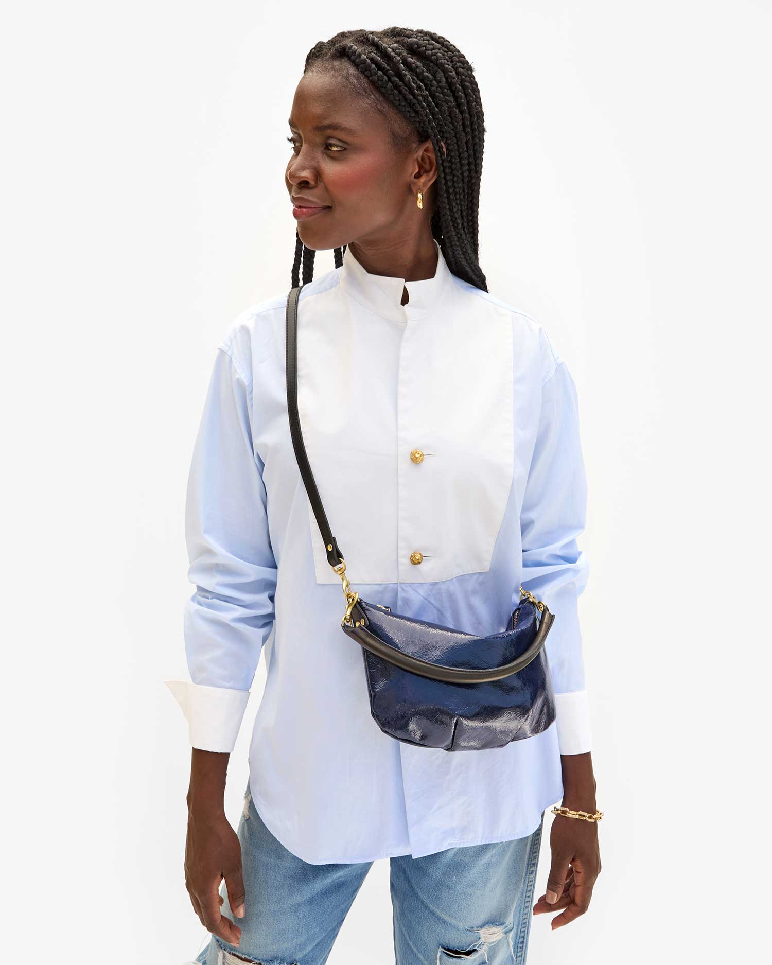 A person with braided hair stands looking to the side, wearing a light blue and white button-up shirt, distressed jeans, and Clare Vivier&#39;s Petite Moyen S24 mini messenger bag with crossbody straps. The shirt features a high collar and gold buttons against a plain white background.