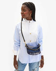 A person with braided hair stands looking to the side, wearing a light blue and white button-up shirt, distressed jeans, and Clare Vivier's Petite Moyen S24 mini messenger bag with crossbody straps. The shirt features a high collar and gold buttons against a plain white background.