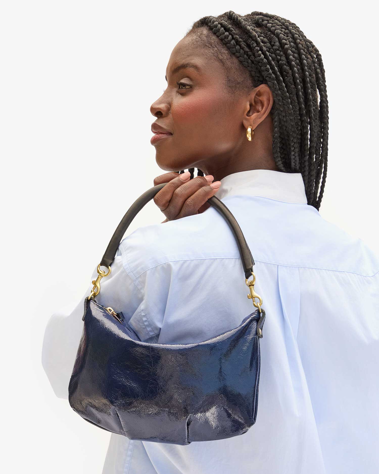 A person with braided hair is turned sideways, showcasing a shiny, dark blue Petite Moyen S24 mini messenger bag by Clare Vivier over their shoulder. They are wearing a light blue shirt and gold hoop earrings while gazing slightly upwards against a plain background.