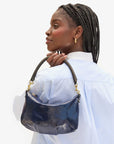 A person with braided hair is turned sideways, showcasing a shiny, dark blue Petite Moyen S24 mini messenger bag by Clare Vivier over their shoulder. They are wearing a light blue shirt and gold hoop earrings while gazing slightly upwards against a plain background.