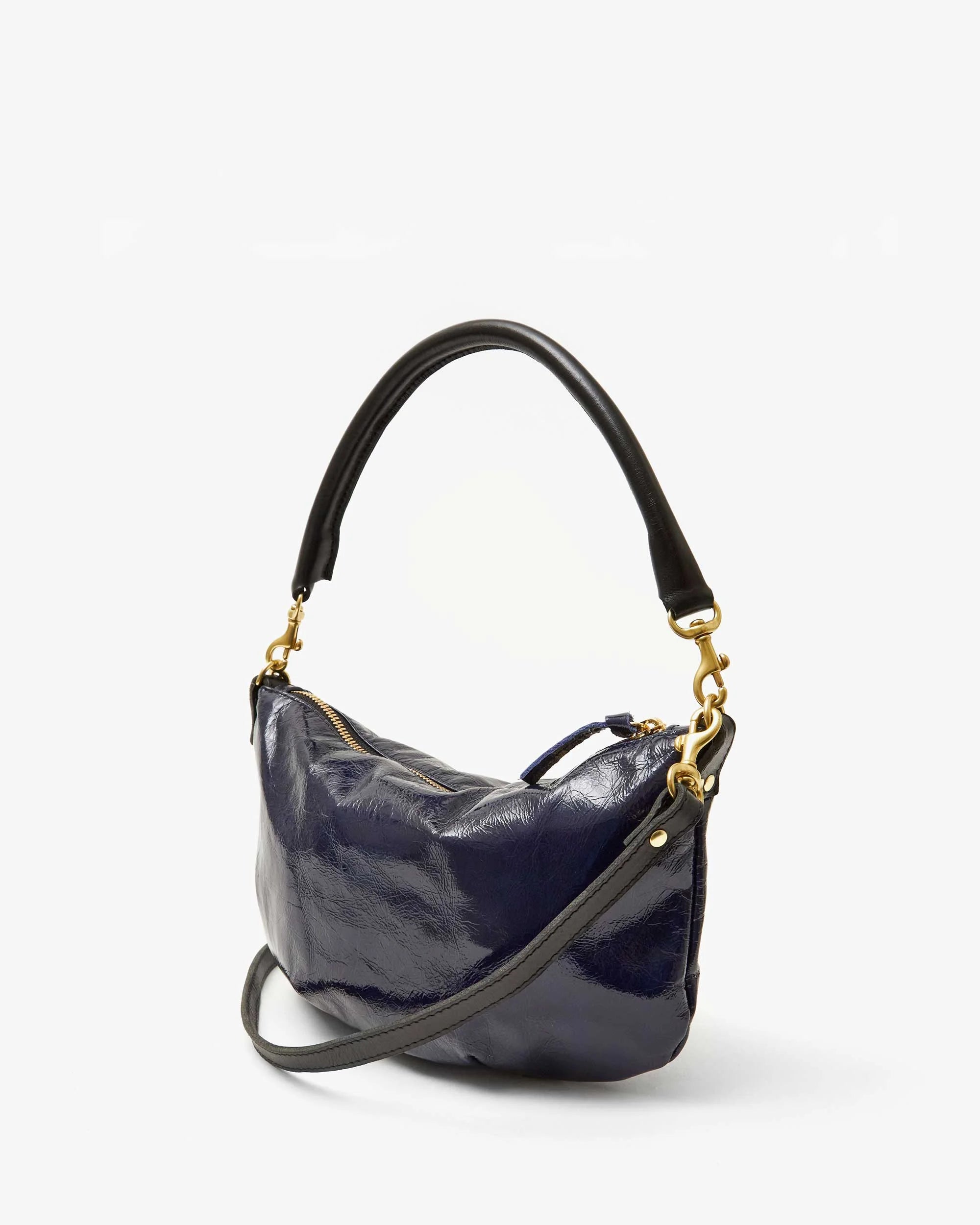 The Petite Moyen S24 by Clare Vivier is a shiny dark blue leather handbag with a soft texture. It features a curved black handle and striking gold hardware, which make it really stand out. Its crossbody straps add versatility, while the plain white background highlights the bag&#39;s elegance.