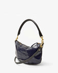 The Petite Moyen S24 by Clare Vivier is a shiny dark blue leather handbag with a soft texture. It features a curved black handle and striking gold hardware, which make it really stand out. Its crossbody straps add versatility, while the plain white background highlights the bag's elegance.