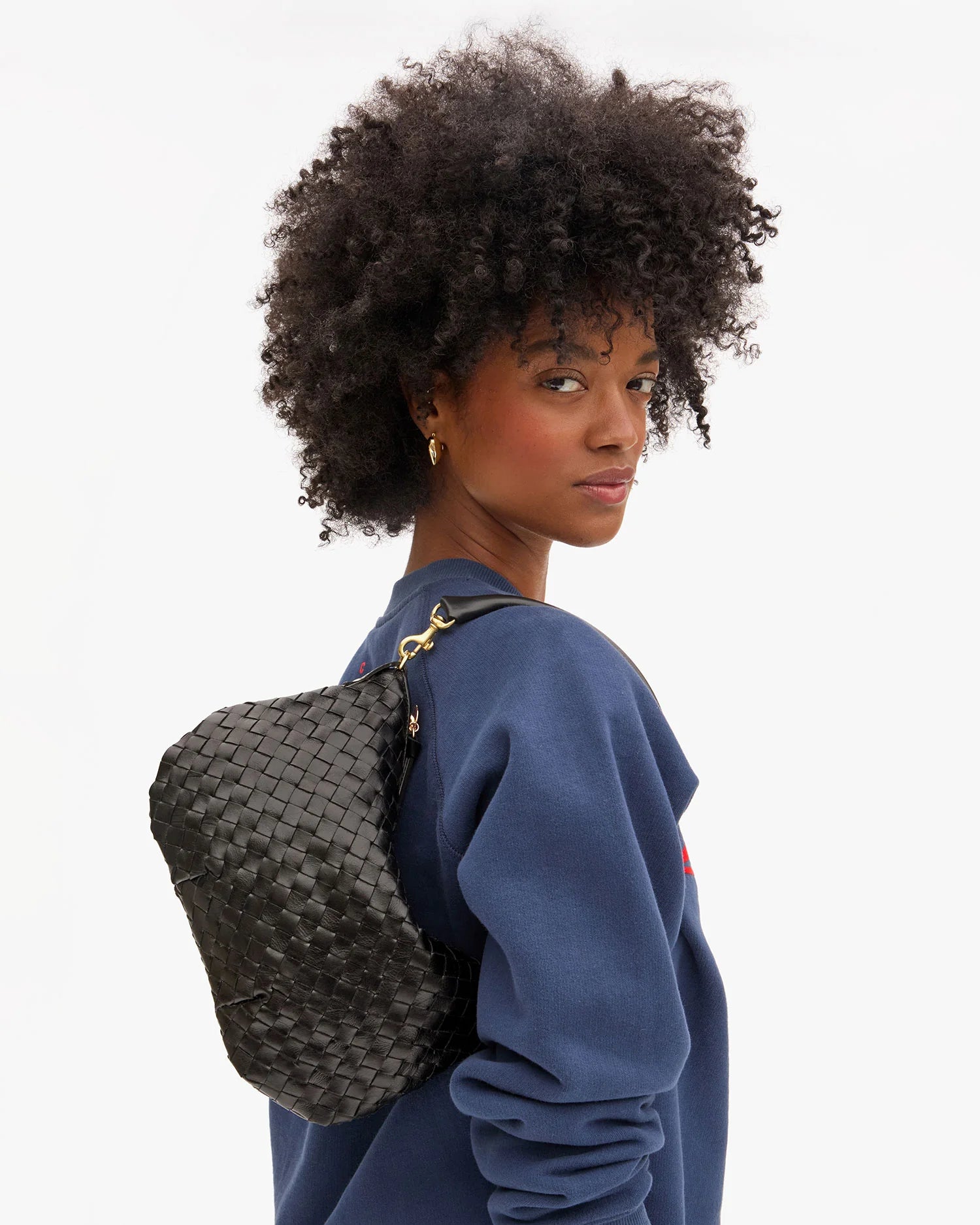 A woman with curly hair in a blue sweater looks over her shoulder, carrying the Clare Vivier Petit Moyen Messenger S25, a black handwoven leather mini crossbody bag. The background is plain white.