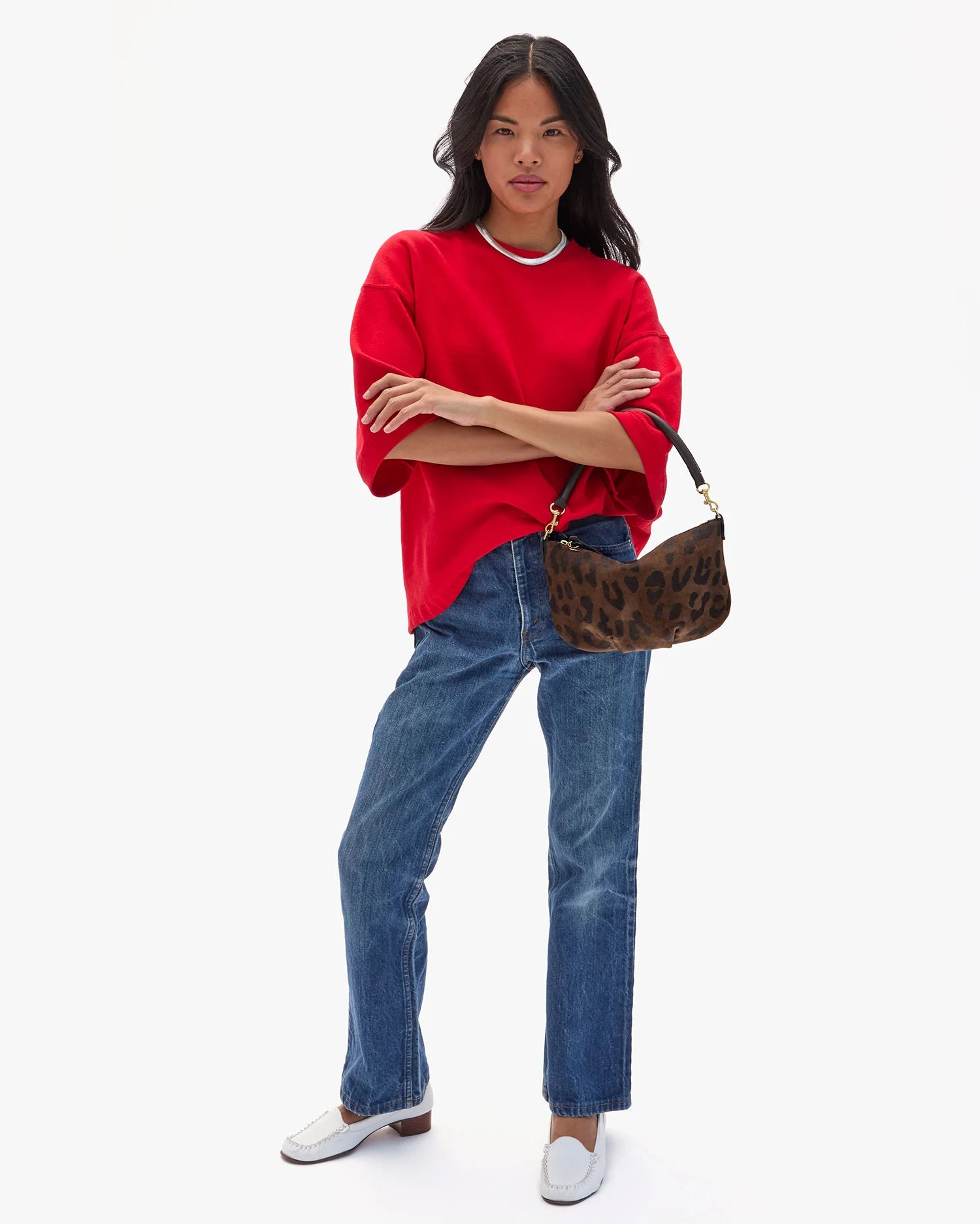 A person with long dark hair in a loose red T-shirt, blue jeans, and white shoes stands confidently holding the Clare Vivier Petit Moyen Messenger S25 with crossbody straps against a plain white background.