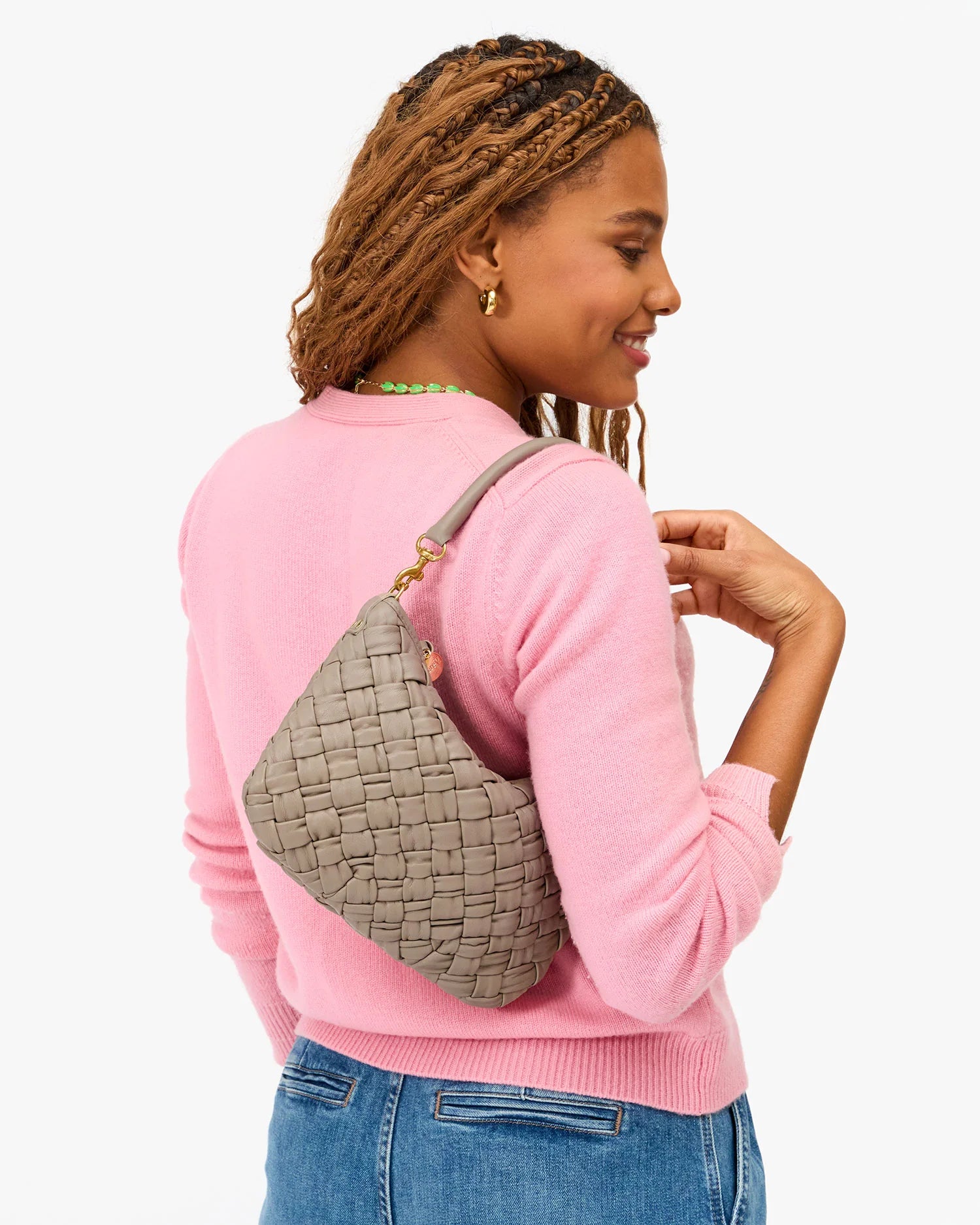 A woman with braided hair, in a pink sweater and blue jeans, smiles while holding the Clare Vivier Petite Moyen S25 mini messenger bag with crossbody straps over her shoulder against a white background.