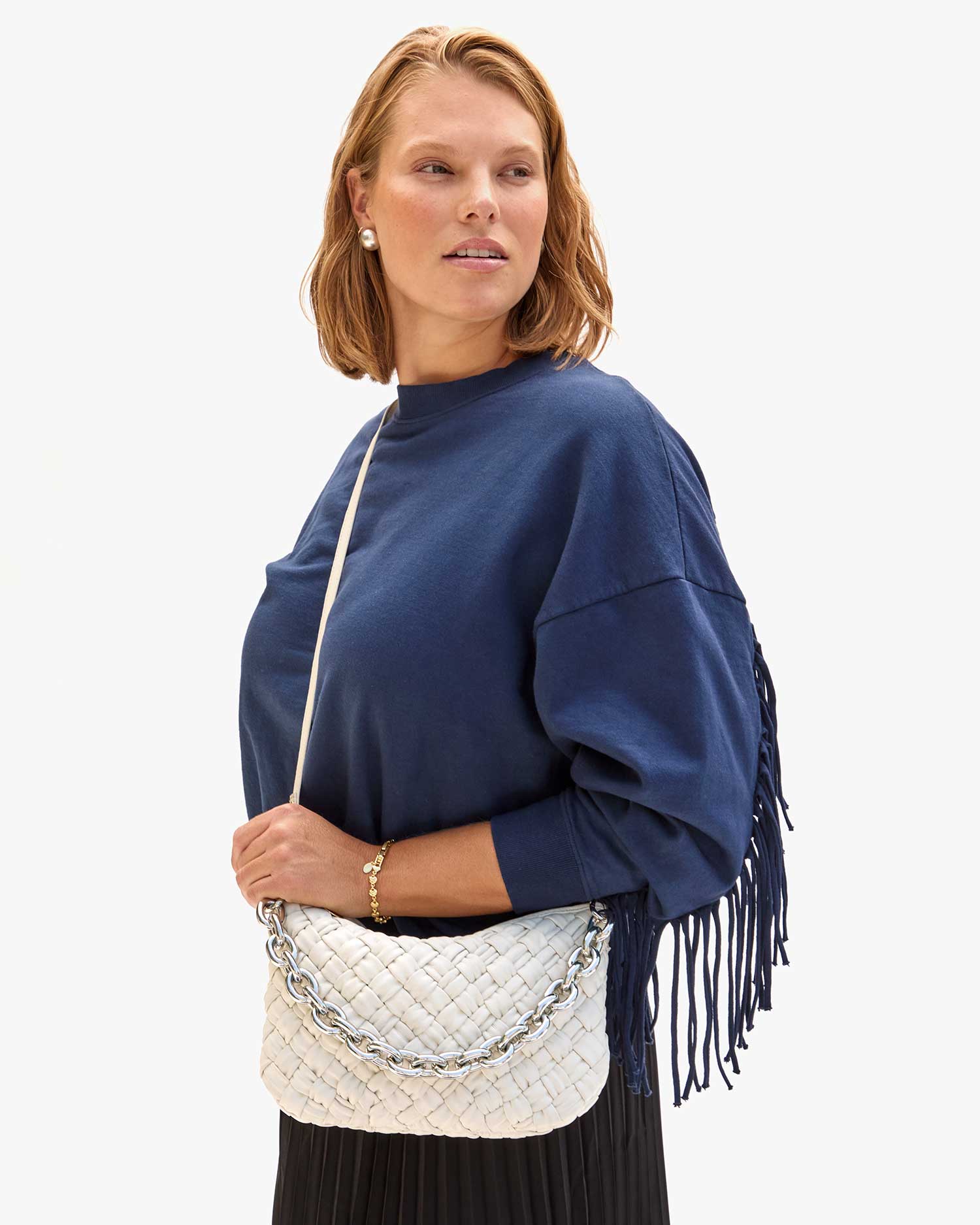 Standing against a plain background, a person with shoulder-length hair wears a blue fringe sweater and a black pleated skirt, holding the Clare Vivier Petite Moyen S24—an elegant white handwoven leather handbag featuring a silver chain strap. They are looking to the side.