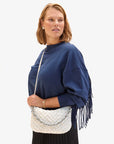 Standing against a plain background, a person with shoulder-length hair wears a blue fringe sweater and a black pleated skirt, holding the Clare Vivier Petite Moyen S24—an elegant white handwoven leather handbag featuring a silver chain strap. They are looking to the side.