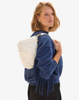 A person wearing a blue top and pleated skirt carries the Clare Vivier Petite Moyen S24, a white handwoven leather bag with fringe detail, over one shoulder. They stand against a plain background, showcasing the elegance of its tubular straps.