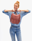 A smiling woman in light denim holds the Clare Vivier Petit Moyen Messenger Suede Fringe bag in pink with both hands, standing against a plain white background.