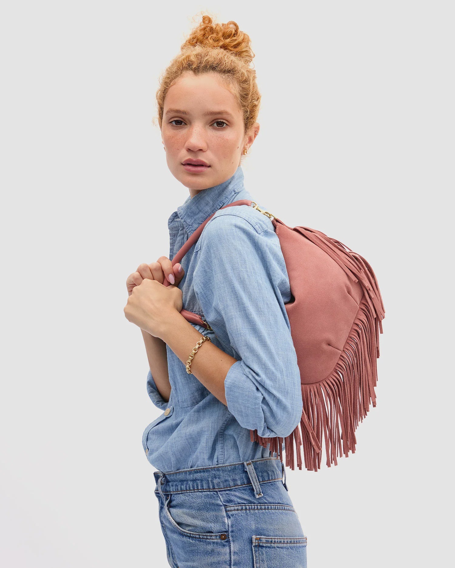 A woman with curly red hair in a bun, wearing a light blue denim shirt and jeans, stands against a plain background with the Clare Vivier Petit Moyen Messenger Suede Fringe bag in mauve suede over her shoulder.