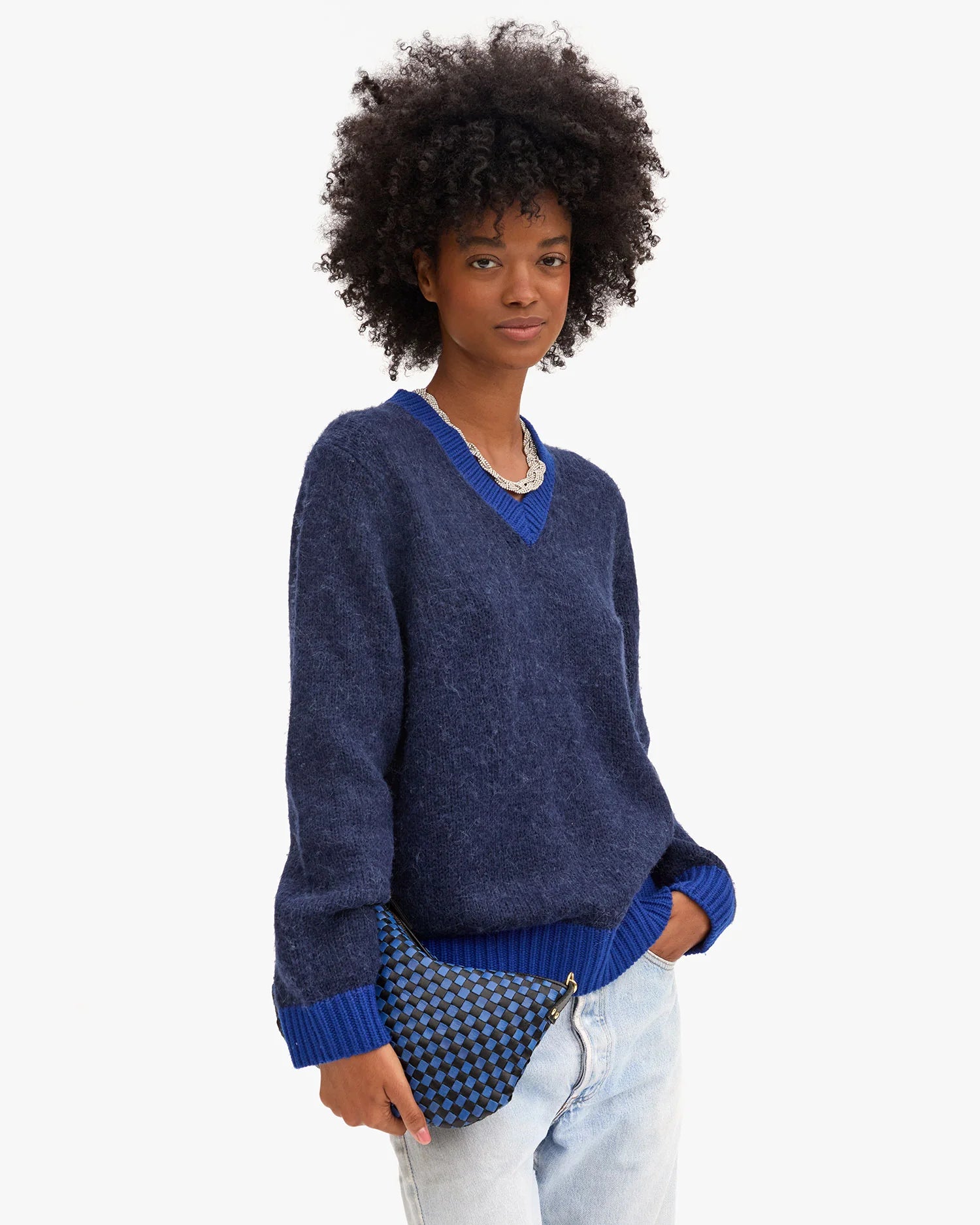 A person with short curly hair wears a loose-fitting navy V-neck sweater and light-wash jeans, carrying the Clare Vivier Petit Moyen Messenger S25 handwoven leather mini bag with crossbody straps against a plain white background.
