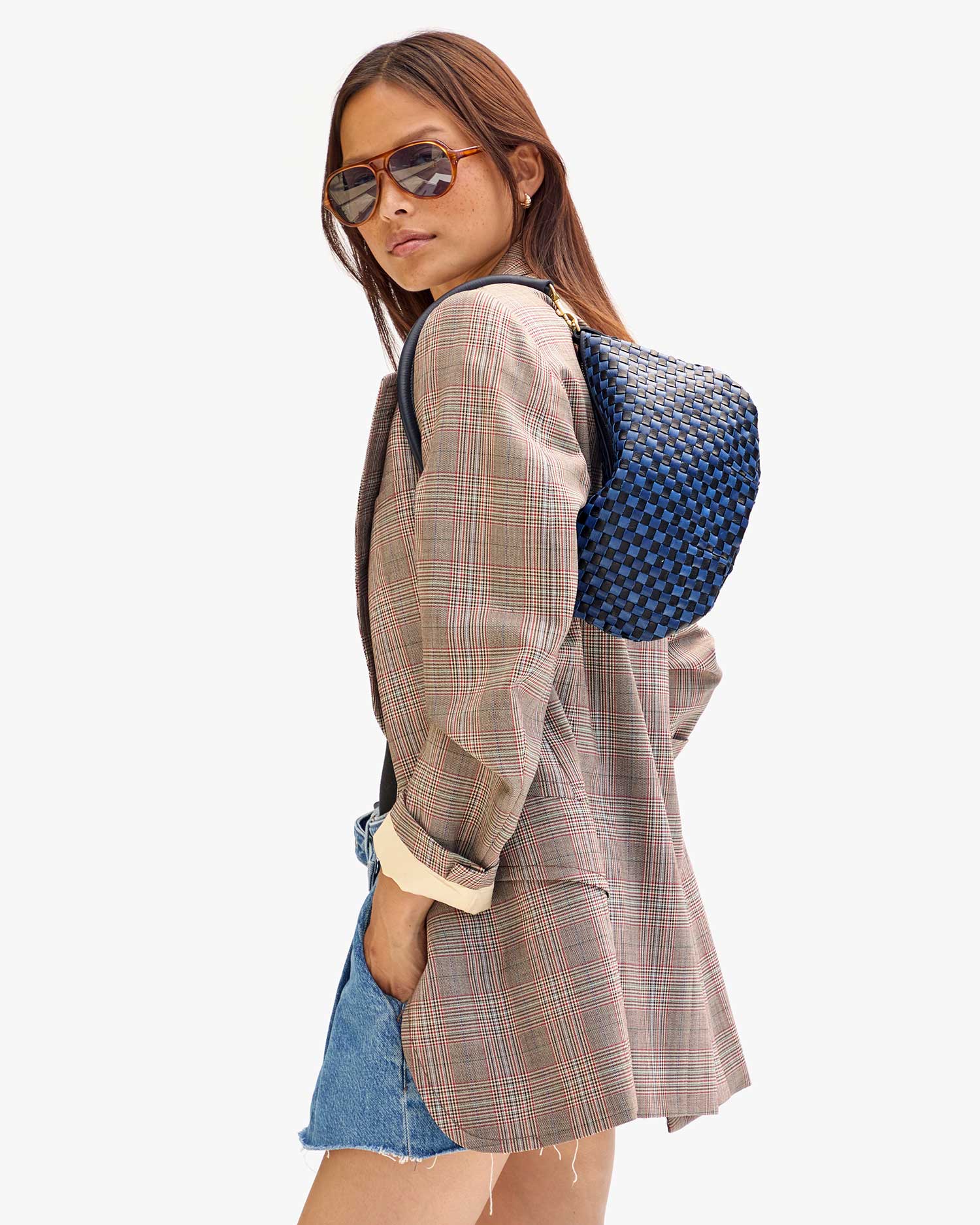 A person dressed in sunglasses, a plaid blazer, denim shorts, and a Clare Vivier Petite Moyen S24 backpack featuring crossbody straps stands against a plain background. They glance over their shoulder with one hand tucked into the blazer pocket.