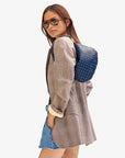 A person dressed in sunglasses, a plaid blazer, denim shorts, and a Clare Vivier Petite Moyen S24 backpack featuring crossbody straps stands against a plain background. They glance over their shoulder with one hand tucked into the blazer pocket.