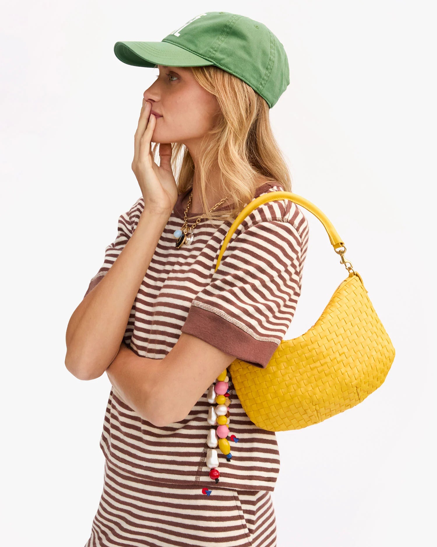A woman in a green baseball cap and brown striped shirt stands in profile, hand on her chin, carrying the Clare Vivier Petit Moyen Messenger S25—a yellow woven crossbody accented with colorful beads—over her shoulder.
