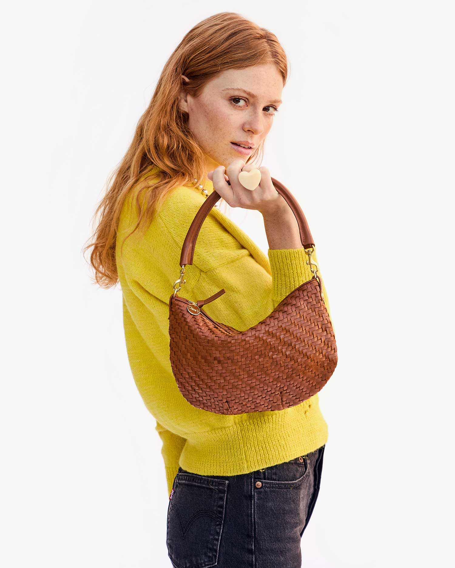 A person with long red hair, wearing a yellow sweater and dark jeans, holds the Clare Vivier Petite Moyen handwoven leather handbag. They glance over their shoulder against a plain white backdrop.