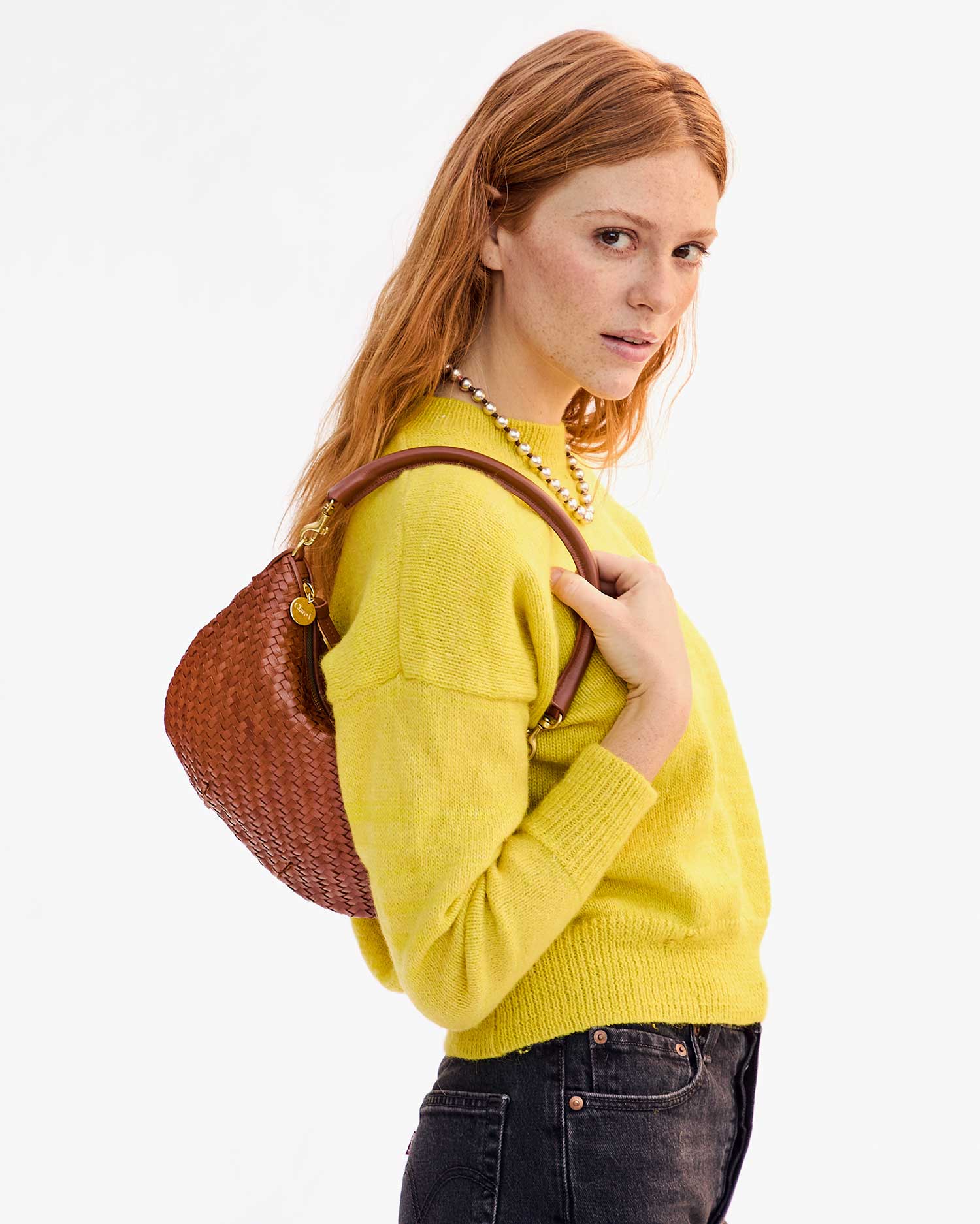 A woman with long red hair models her style with the Clare Vivier Petite Moyen handwoven leather handbag over her shoulder. Her yellow sweater and black pants complement her pearl necklace, radiating elegance against a white backdrop.