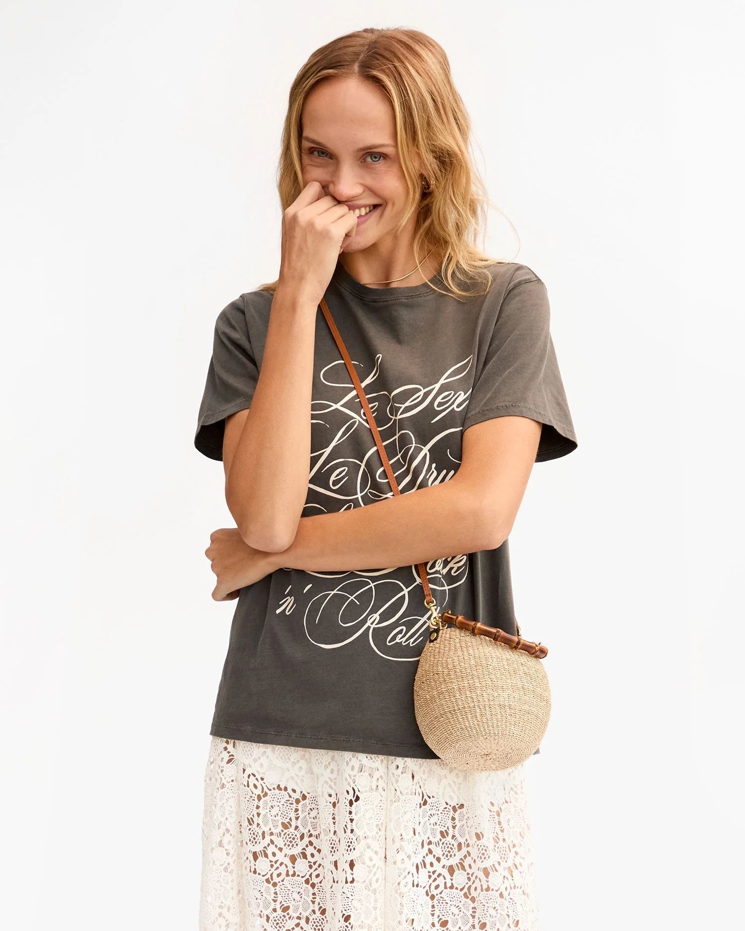 A smiling woman with wavy blonde hair wears a dark graphic t-shirt, a white lace skirt, and carries the Petite Pot de Miel straw bag by Clare Vivier. She stands against a plain light background, one hand gently touching her face.