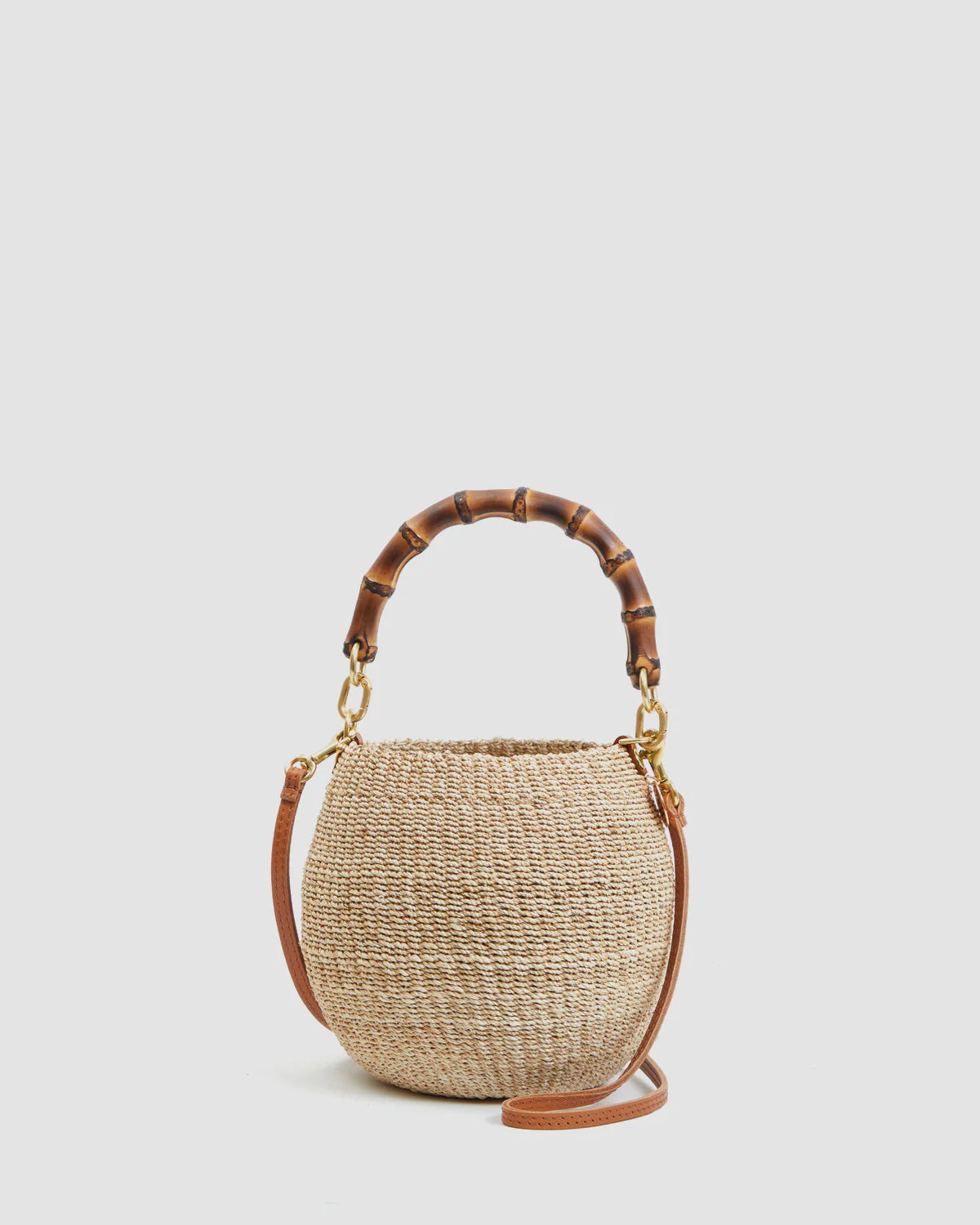 The Clare Vivier Petite Pot de Miel is a round, handwoven straw basket bag featuring a curved bamboo handle and a detachable brown leather shoulder strap, shown against a plain light background.