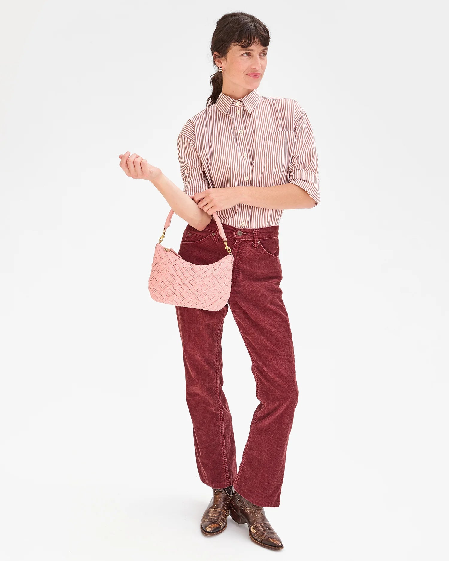 A person with long dark hair, dressed in a striped shirt and red pants, confidently holds the Petite Moyen by Clare Vivier—a pink woven handbag with crossbody straps. They glance to the side with one hand on their hip, completing the look with brown shoes.