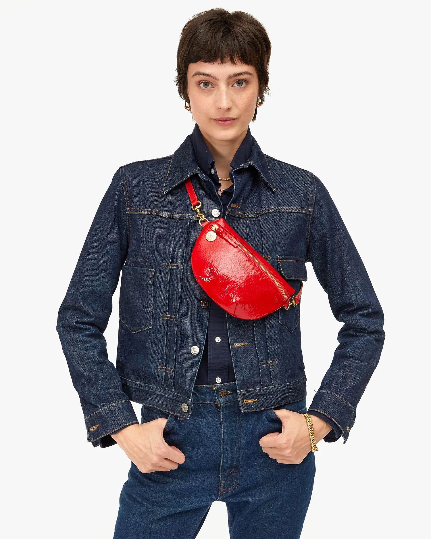 A person in a dark denim jacket and jeans poses confidently with hands in pockets. They have short brown hair and wear the Clare Vivier Petit Fanny F25, an adjustable petite fanny pack, across their chest. The background is plain white.