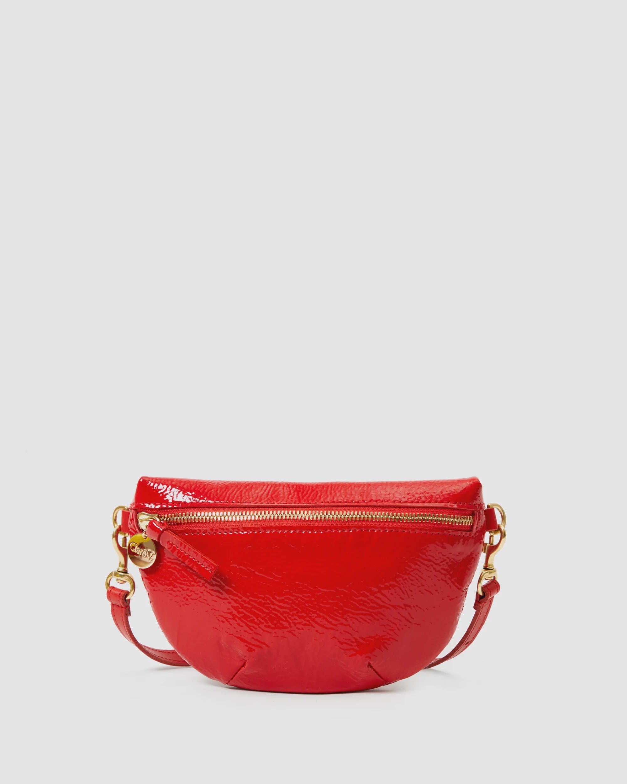 The Clare Vivier Petit Fanny F25 is a shiny, bright red patent leather crossbody bag with a textured finish, gold-tone zipper and hardware, rounded shape, and detachable strap, shown against a plain light gray background.