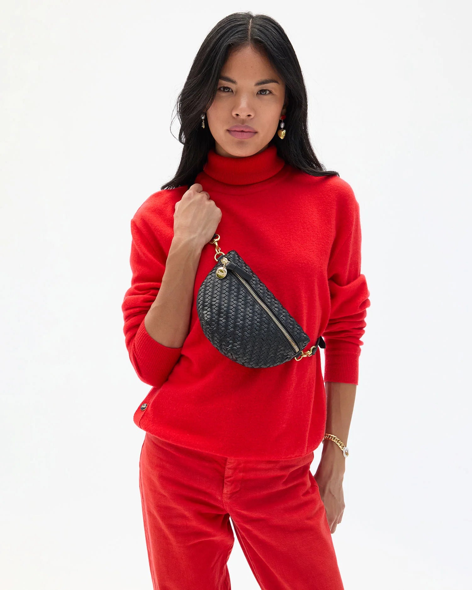 A woman with long black hair wears a bright red turtleneck and pants, carrying the Clare Vivier Petit Fanny F25 textured patent leather crossbody bag with adjustable strap over her shoulder. The background is plain white.