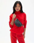 A woman with long black hair wears a bright red turtleneck and pants, carrying the Clare Vivier Petit Fanny F25 textured patent leather crossbody bag with adjustable strap over her shoulder. The background is plain white.